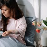 A woman sits in bed enjoying breakfast with milk and flowers nearby, bathed in morning light.